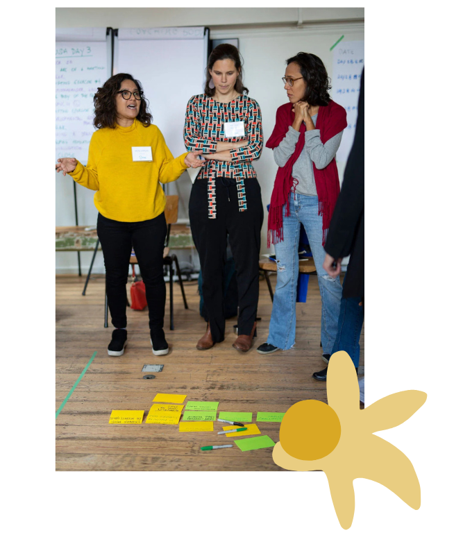 Three people in brightly coloured clothing stand around post-its on the ground as one speaks emphatically. Spring facilitation training in Amsterdam 2019.