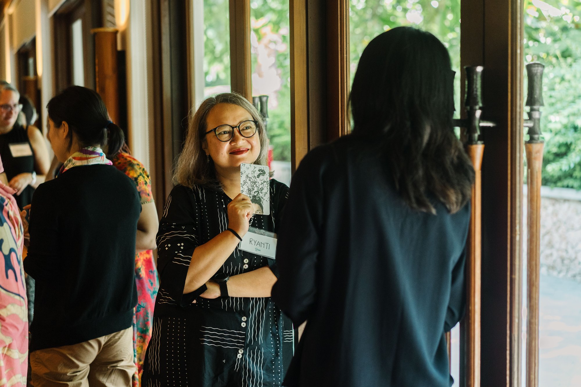 A woman in a black shirt with white stripes leans on a glass window in deep conversation with another participant of the FIRE Program in Bali, Indonesia.