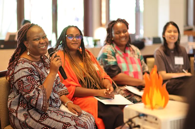 FIRE Participants share during an in-person training in Bali, Indonesia. A woman wears auburn dreadlocks and a darkly patterned dress, smiling as she speaks into a microphone.